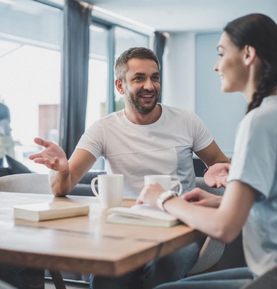 happy adult man gesturing and talking with girlfriend at table with coffee cups and books at home happy adult man gesturing and talking with girlfriend at table with coffee cups and books at home